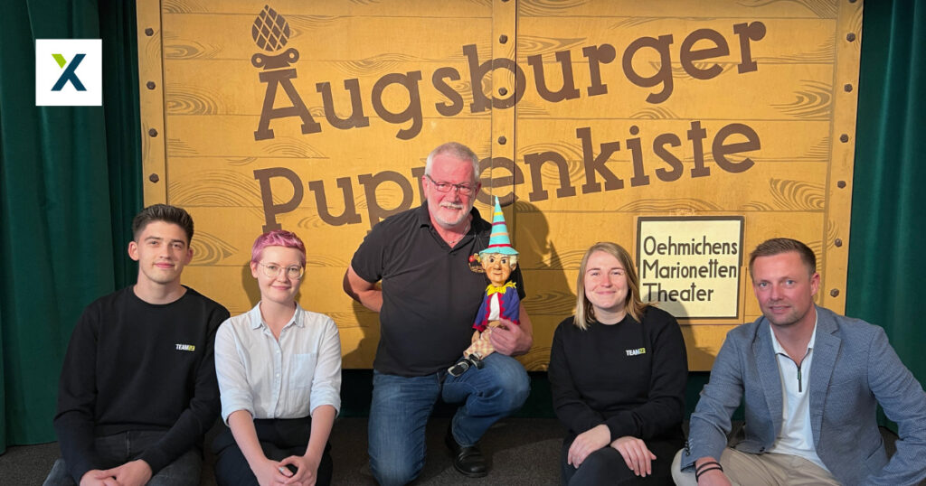 Eine Gruppe von fünf Menschen posiert vor einem Schild mit der Aufschrift „Augsburger Puppenkiste“.