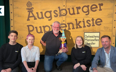Eine Gruppe von fünf Menschen posiert vor einem Schild mit der Aufschrift „Augsburger Puppenkiste“.
